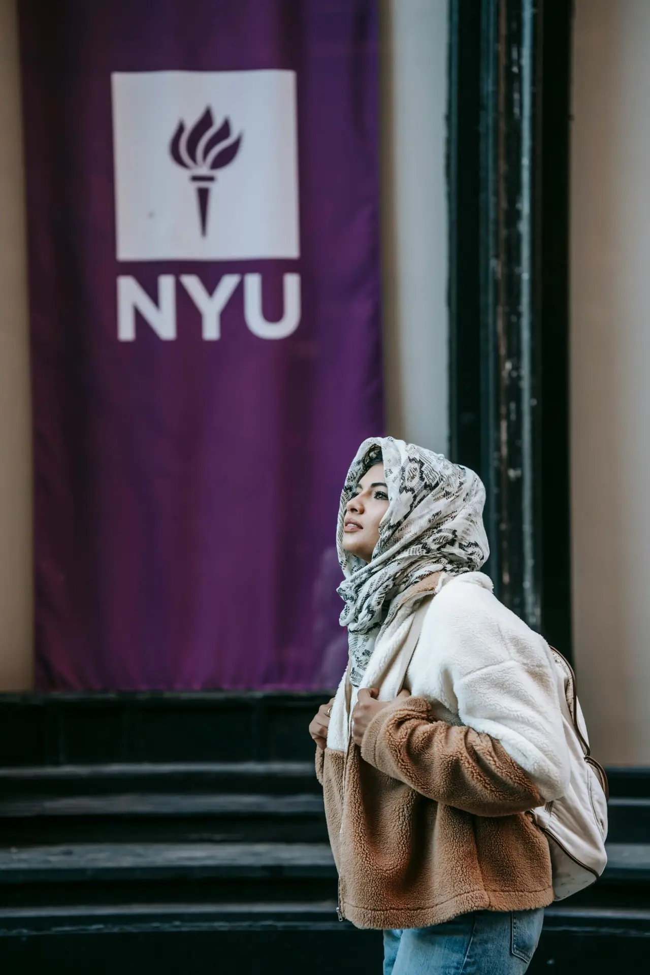 Young Muslim woman walking by NYU with backpack and hijab, embracing independent student life.